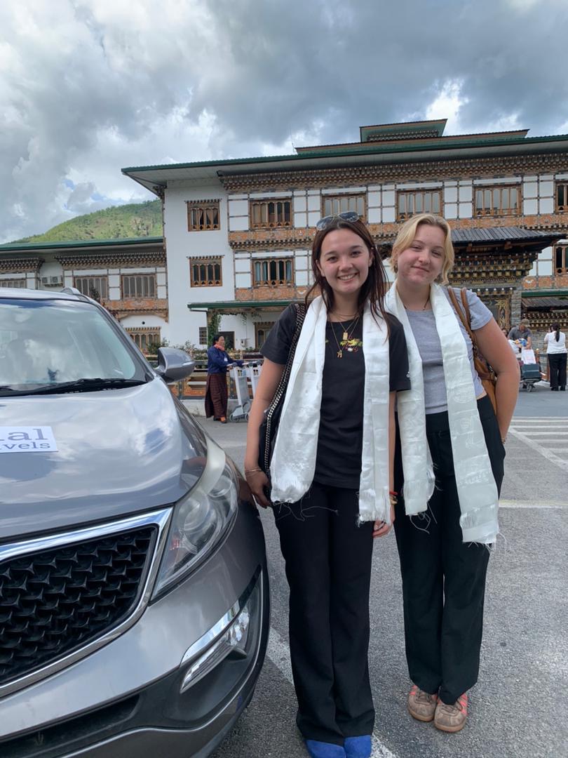 Two young women wearing white ceremonial scarves stand beside a grey vehicle in front of a traditional Bhutanese building, under a cloudy sky.