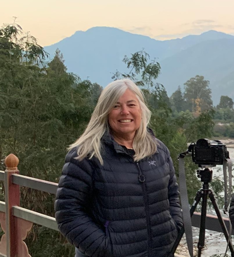 A woman with long white hair wearing a black puffer jacket smiles at the camera, standing on a bridge with a camera tripod beside her and mountains in the background.