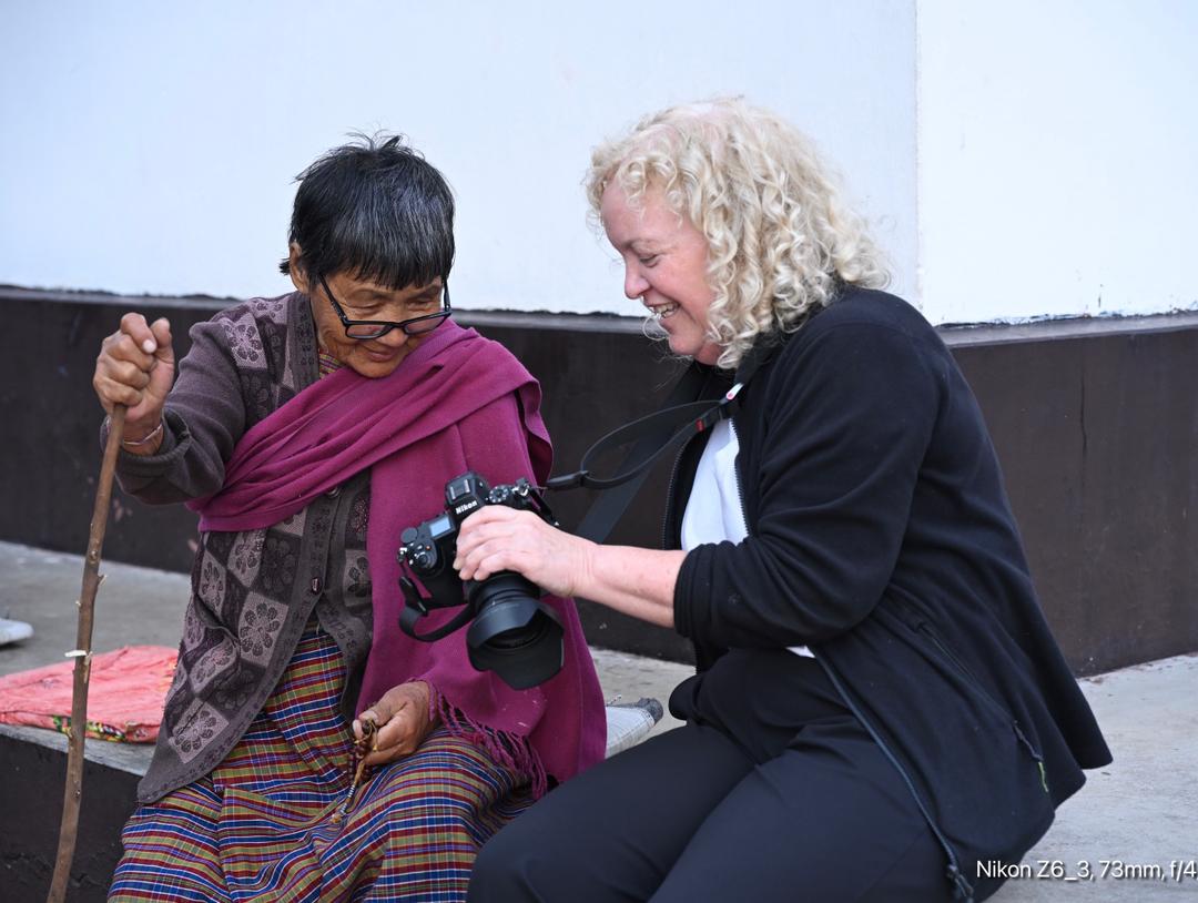 A woman with curly hair and a camera engages in a friendly conversation with an elderly woman, who is holding a stick and wearing traditional clothing. They are sitting outdoors with a plain wall in the background.